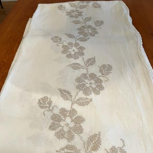 May include: A white linen tablecloth with a delicate floral pattern embroidered in a light brown color. The tablecloth has a scalloped edge.