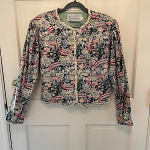 May include: A floral print jacket with a round neckline and long sleeves. The jacket features a pattern of flowers, fruits, and foliage in shades of pink, green, and white. It has a button-up front and a decorative trim along the edges.