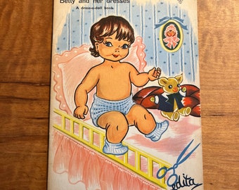May include: A vintage dress-up doll book featuring a cartoon illustration of a little girl in a blue diaper sitting on a bed with a teddy bear. The book cover has the title "Betty and her dresses" and the text "A dress-a-doll book".