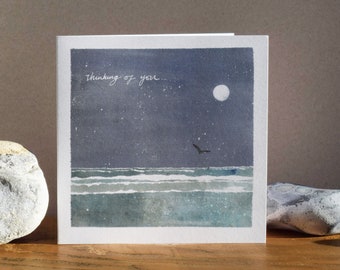 May include: A watercolor card with a night sky scene. The sky is a dark blue with white stars and a full moon. The ocean is a dark blue with white waves. A single bird flies over the ocean. The card says "thinking of you..." in white script.