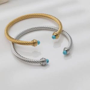 May include: Two open cuff bracelets featuring a twisted cable design. One bracelet is gold-toned and the other is silver-toned. Each end of both bracelets is capped with a smooth, round, bright turquoise stone set in a polished metal housing.