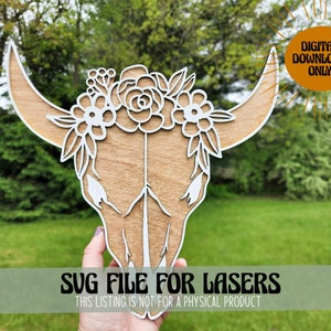 May include: A wooden cutout of a bull skull with a floral wreath around the horns. The skull is light brown and the flowers are white. The text "SVG FILE FOR LASERS" and "THIS LISTING IS NOT FOR A PHYSICAL PRODUCT" is at the bottom of the image.