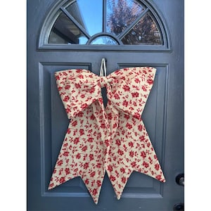 May include: A large, decorative bow made of cream-colored fabric with a red floral pattern, hanging on a blue door. The bow is tied in the center and has long, pointed tails. The door has a window at the top.