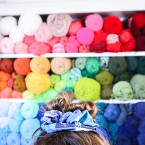 K&ouml;nnte beinhalten: Eine Person mit braunen Haaren tr&auml;gt ein blaues und wei&szlig; gemustertes Haargummi im Haar. Der Hintergrund ist eine Wand mit Regalen, die mit bunten Garnen gef&uuml;llt sind.