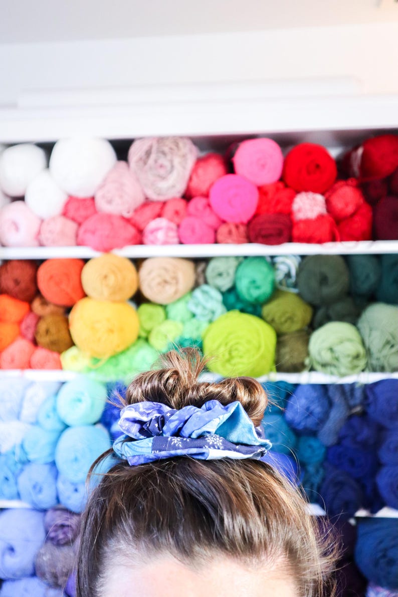K&ouml;nnte beinhalten: Eine Person mit braunen Haaren tr&auml;gt ein blaues und wei&szlig; gemustertes Haargummi im Haar. Der Hintergrund ist eine Wand mit Regalen, die mit bunten Garnen gef&uuml;llt sind.