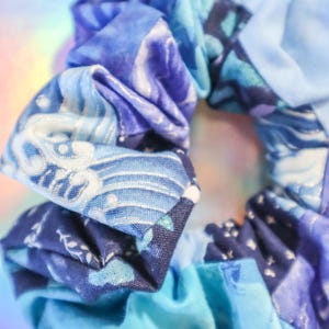 K&ouml;nnte beinhalten: Ein blauer und wei&szlig;er gemusterter Haargummi aus Stoff. Der Haargummi besteht aus mehreren zusammengen&auml;hten Stoffteilen.