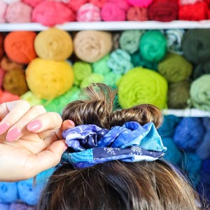 K&ouml;nnte beinhalten: Nahaufnahme von Haaren, die mit einem blauen und wei&szlig;en gemusterten Haargummi zu einem Dutt gebunden sind. Der Hintergrund ist eine Wand mit bunten Wollkn&auml;ueln.