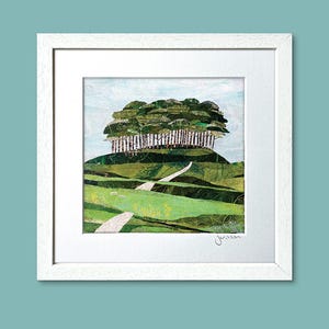 May include: Framed art print of a collage landscape. The artwork shows a green hill with trees and a winding path. The sky is light blue. The artwork is in a white frame. The artist's signature is visible.