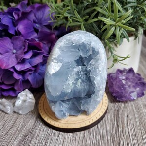 May include: A blue celestite crystal cluster with a natural, raw, and rough texture. The crystal is on a wooden stand with a circular design.