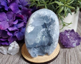 May include: A blue celestite crystal cluster with a natural, raw, and rough texture. The crystal is on a wooden stand with a circular design.