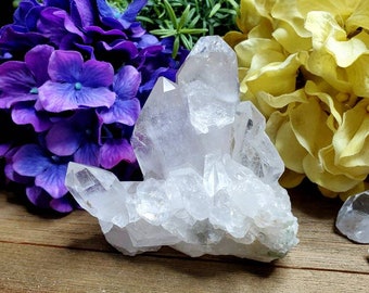 May include: A large, clear quartz crystal cluster with multiple points. The crystal is sitting on a wooden surface with purple and yellow flowers in the background.