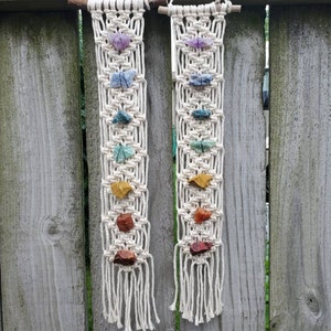 May include: Two macrame wall hangings with colorful stones. Each hanging features a white macrame design with seven stones in a rainbow color pattern.