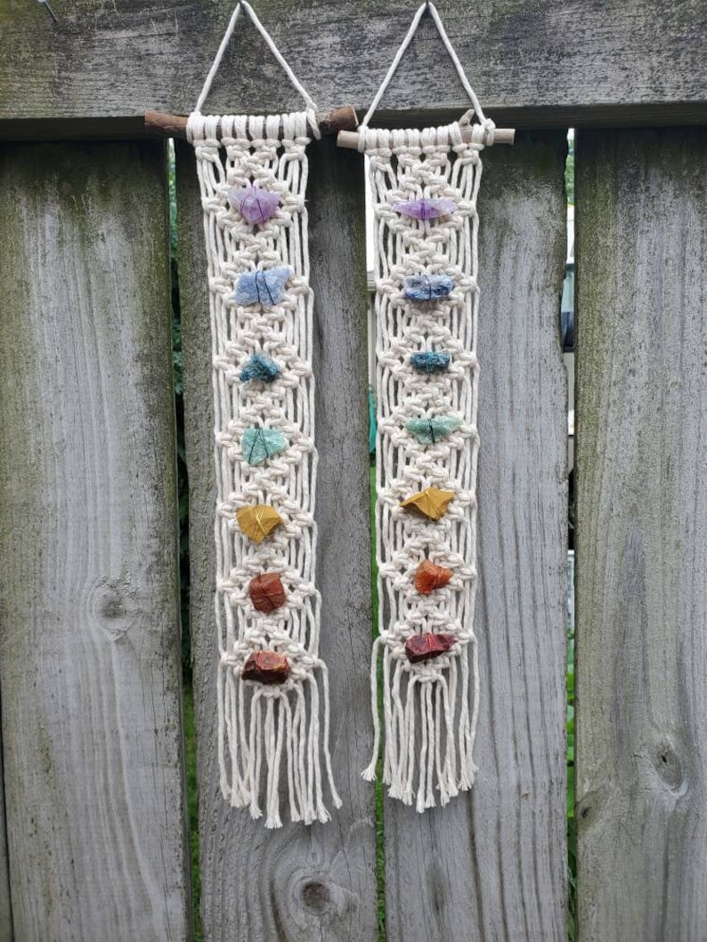 May include: Two macrame wall hangings with colorful stones. Each hanging features a white macrame design with seven stones in a rainbow color pattern.