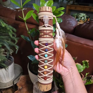 May include: A cylindrical wooden staff wrapped in brown suede leather with a crisscross pattern of light beige cord. The staff features a clear quartz point at the top and seven small, colorful spherical beads arranged vertically in a row.
