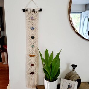 May include: A macrame wall hanging with a wooden dowel at the top. The macrame is cream colored and has a geometric pattern. There are seven small stones of various colors and shapes hanging from the macrame. The wall hanging is hanging on a white wall. A green plant in a white pot is on a table in front of the wall hanging.