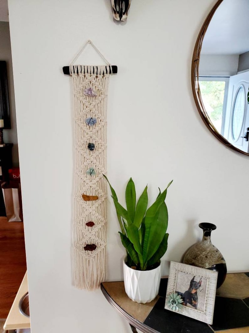 May include: A macrame wall hanging with a wooden dowel at the top. The macrame is cream colored and has a geometric pattern. There are seven small stones of various colors and shapes hanging from the macrame. The wall hanging is hanging on a white wall. A green plant in a white pot is on a table in front of the wall hanging.