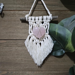 May include: A white macrame wall hanging with a pink heart-shaped crystal in the centre. The hanging is attached to a wooden branch and has a fringe border.
