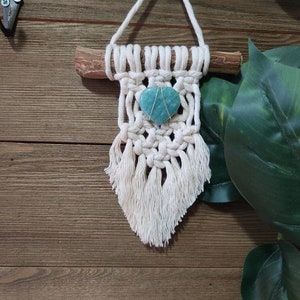 May include: A white macrame wall hanging with a light blue heart-shaped stone in the centre. The hanging is attached to a wooden dowel and has a fringe at the bottom.