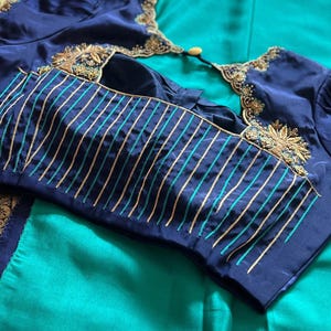 May include: Navy blue blouse with gold embroidery and turquoise and gold striped sleeves. The blouse is on a teal green fabric. The blouse has a round neckline and short sleeves. The blouse is made of a silky material.