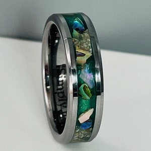 May include: A silver ring with a green band and embedded seashells. The band is beveled and has a brushed finish. The inscription "Simply" is visible on the side of the ring.