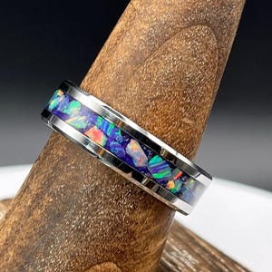 May include: A silver ring with a band inlaid with a mosaic of colorful opal fragments. The opal features shades of blue, green, purple, and orange. The ring is displayed on a wooden ring holder, showcasing its design and craftsmanship.