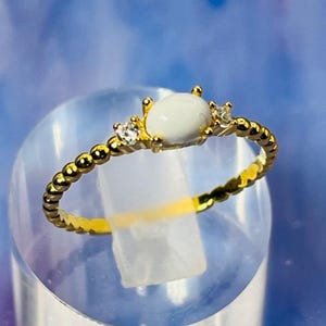 Può includere: Un anello color oro con una pietra ovale bianca al centro, affiancata da due pietre trasparenti più piccole. La fascia presenta un design strutturato a perline. L'anello è esposto su un supporto cilindrico trasparente su sfondo blu.