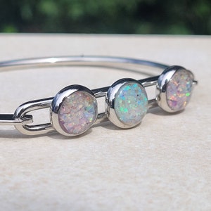 May include: A silver bangle bracelet with three round opal-like stones. The stones are white with iridescent blue and pink flecks.