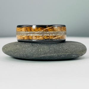 May include: A black tungsten carbide ring with a wood inlay. The wood inlay is a light brown color with white streaks. The ring is sitting on a gray rock.