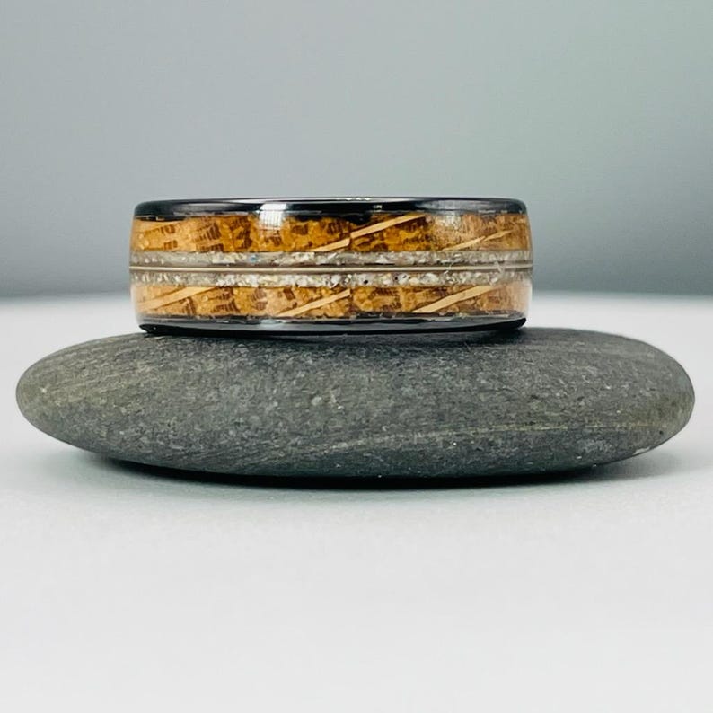 May include: A black tungsten carbide ring with a wood inlay. The wood inlay is a light brown color with white streaks. The ring is sitting on a gray rock.