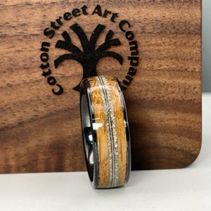 May include: A black tungsten carbide ring with a wood inlay. The wood is a light brown color with white streaks. The ring is on a wooden surface.