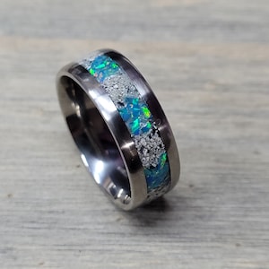 Puede incluir: Un anillo de titanio plateado con una banda de ópalo iridiscente y motas blancas.