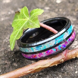 May include: A black tungsten carbide ring with a multi-colored inlay of iridescent opal and pink glitter. The ring is sitting on a branch and a green leaf.
