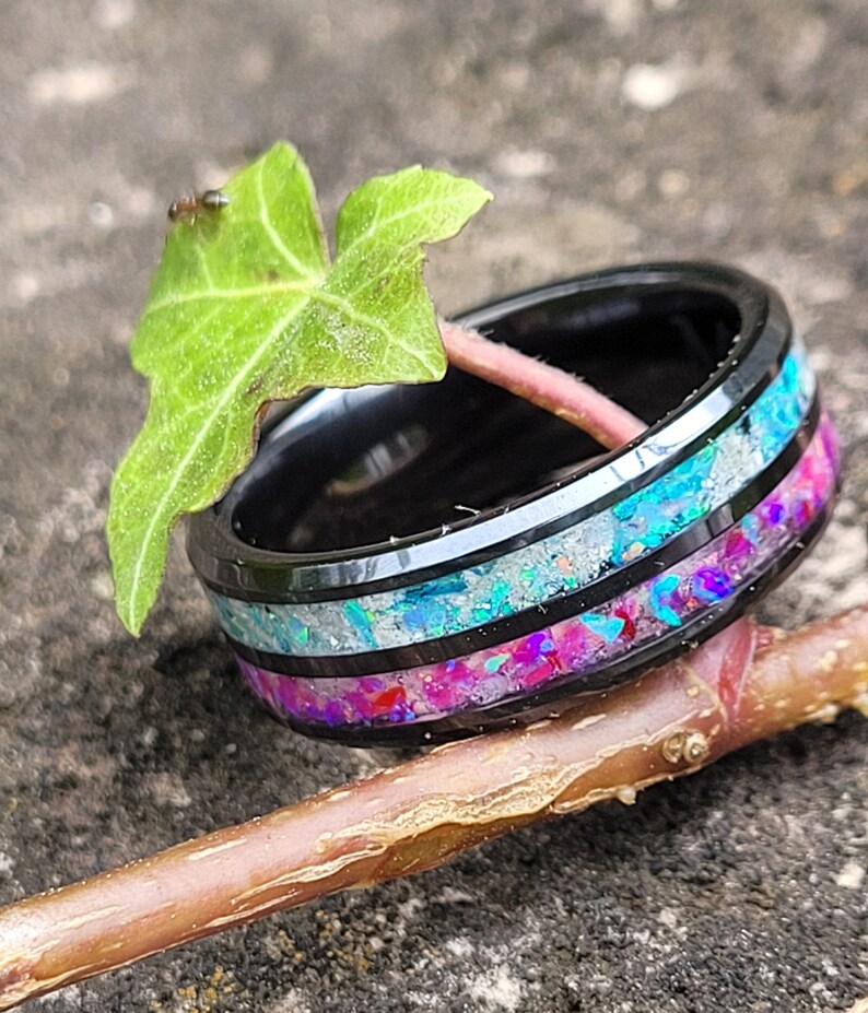 May include: A black tungsten carbide ring with a multi-colored inlay of iridescent opal and pink glitter. The ring is sitting on a branch and a green leaf.