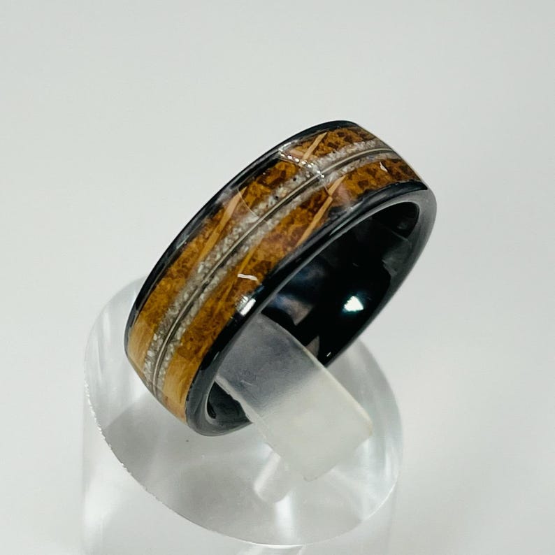 May include: A black ceramic ring with a brown wood inlay and white accents. The ring is on a clear display stand.