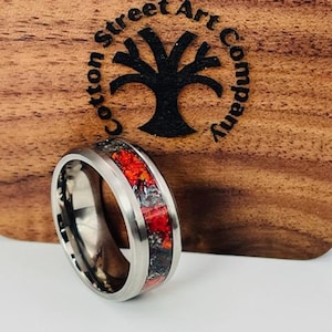 May include: A silver titanium ring with a red and black inlay. The ring is on a wooden surface with the words "Cotton Street Art Company" engraved on it.