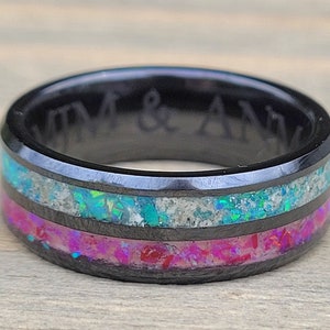 May include: A black tungsten carbide ring with two bands of opal inlay. The top band is a light blue and white opal, and the bottom band is a pink and white opal. The ring is engraved with the text "KIM & ANN".