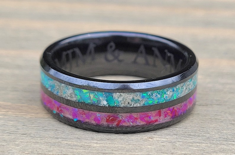 May include: A black tungsten carbide ring with two bands of opal inlay. The top band is a light blue and white opal, and the bottom band is a pink and white opal. The ring is engraved with the text "KIM & ANN".