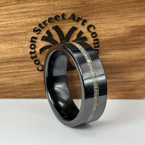 Puede incluir: Un anillo negro con un acabado de metal cepillado y una incrustación central de material de color claro. El anillo está sobre un fondo de madera con el texto "Cotton Street Art Co." visible.