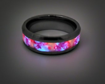 May include: A dark gray ring with a vibrant, glowing inlay. The inlay features a mix of pink, red, and blue colors, creating a striking visual effect. The ring's band appears to be made of a dark metal.
