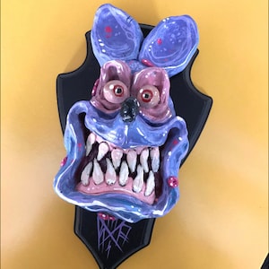 May include: A purple and pink ceramic sculpture of a monster's head with large teeth and eyes. The sculpture is mounted on a black plaque with a silver design.