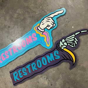 May include: Two wooden signs with the word "RESTROOMS" painted on them. One sign is painted in shades of blue and pink with a hand pointing to the right. The other sign is painted in shades of purple and yellow with a skeleton hand pointing to the right.