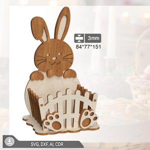 May include: Wooden Easter bunny basket with a light brown finish. The bunny has long ears and is holding a basket with a fence design. Dimensions are 8.4cm x 7.7cm x 15.1cm. The image also includes the text "SVG, DXF, AI, CDR".