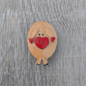 Puede incluir: Un broche de cerdo marrón claro con corazones rojos para las mejillas. El cerdo tiene ojos negros y una nariz negra.