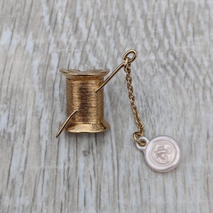 Könnte beinhalten: Eine goldfarbene Garnrolle mit einer Nadel und einem weißen Knopf, der an einer Kette befestigt ist. Der Knopf hat ein Blumenmuster.