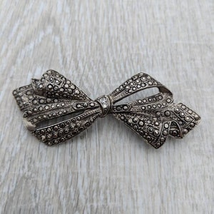 May include: A silver bow brooch with a marbled design and a single clear rhinestone in the centre.