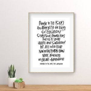 May include: A framed print with black text that reads "Praise to the Lord, the Almighty, the King of Creation!" The print is displayed on a wooden surface with a small potted plant.