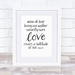 Op de afbeelding: Een ingelijste print met een witte rand toont de woorden "above all, keep loving one another earnestly, since love covers a multitude of sins." De tekst staat in zwart schrift op een witte achtergrond, tegen een witte bakstenen muur.