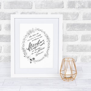 May include: A framed black and white print with floral and arrow accents. The print features script text in Spanish, surrounded by a floral wreath. A gold geometric candle holder sits to the right of the frame.