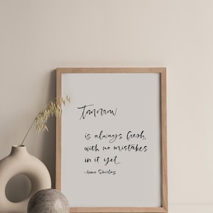 May include: A framed print with the quote "Tomorrow is always fresh, with no mistakes in it yet." in black script. A decorative vase with a sprig of dried grass and a stone sphere are next to the frame. The frame is light brown.