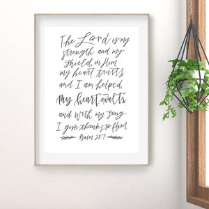 May include: A framed print with the text "The Lord is my strength and my shield, in Him my heart trusts and I am helped, My heart exults and with my song I give thanks to Him. Psalm 28:7" in black script. The frame is light brown.